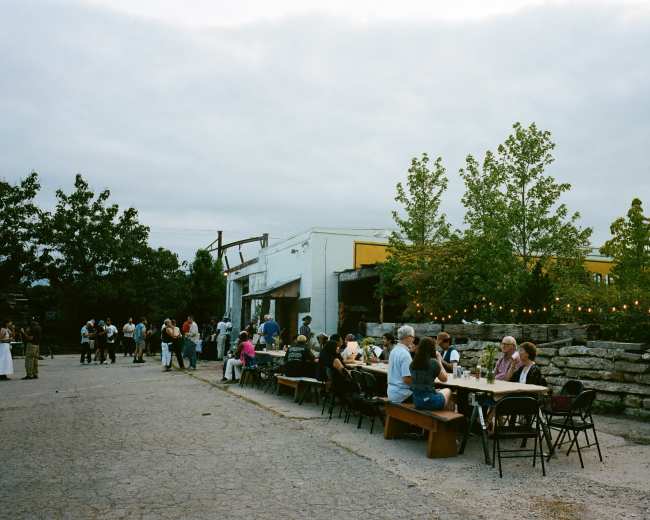 The image shows a gathering of people seated at tables and mingling in an outdoor setting, surrounded by trees and buildings under a cloudy sky.