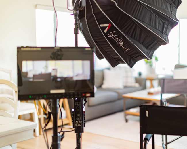 A home filming setup featuring a large softbox light, a monitor with a camera feed, and two empty chairs in a living room setting.
