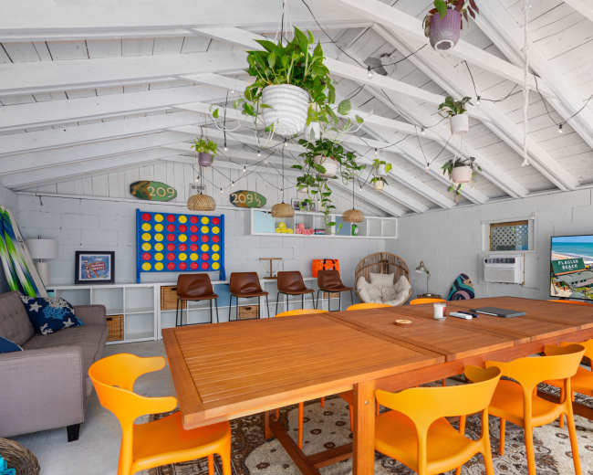 The image shows a bright indoor space with a large wooden table surrounded by orange chairs, a sofa, and shelves adorned with plants and beach-themed decor.