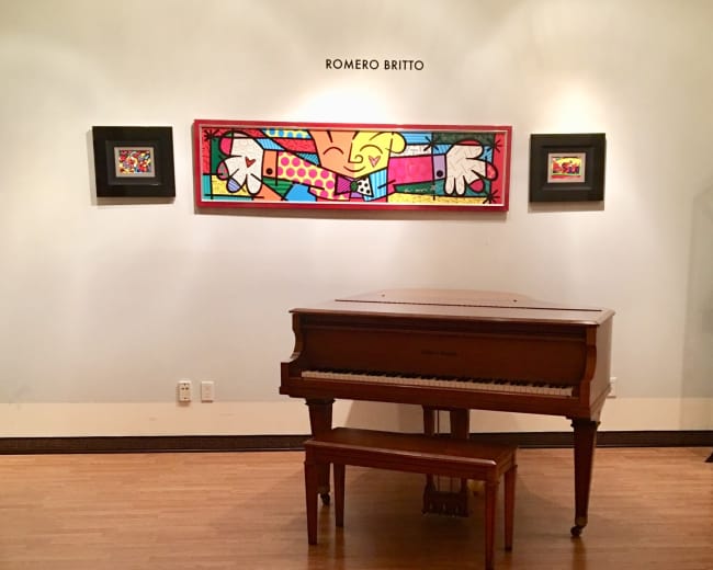 A wooden piano is situated in a gallery beneath a colorful artwork by Romero Britto on the wall.