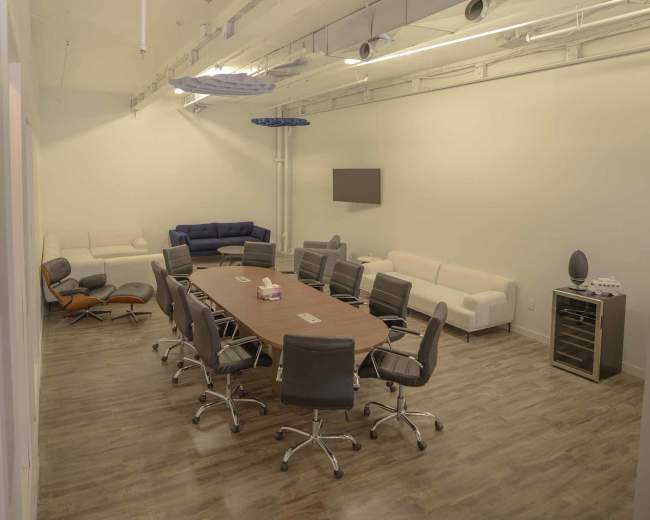 The image shows a modern conference room with a large wooden table surrounded by ergonomic chairs, flanked by white sofas and a wall-mounted television.