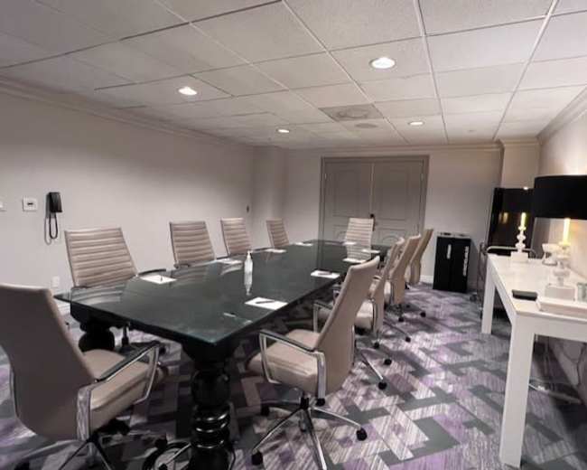 A conference room features a long glass table surrounded by ten ergonomic chairs, with a white side table and modern decor along the walls.