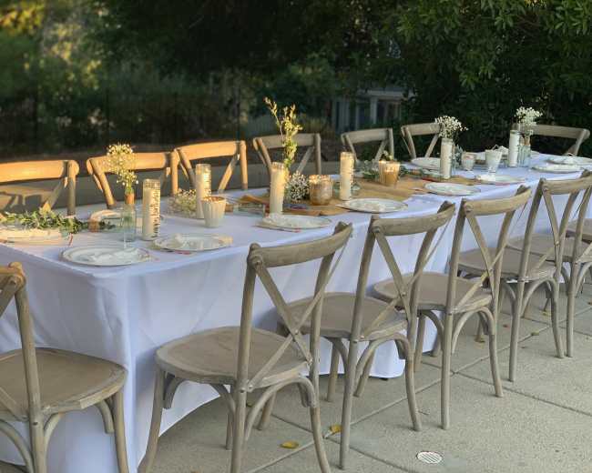A long outdoor dining table is set with white tablecloths, plates, and centerpieces of flowers in jars.