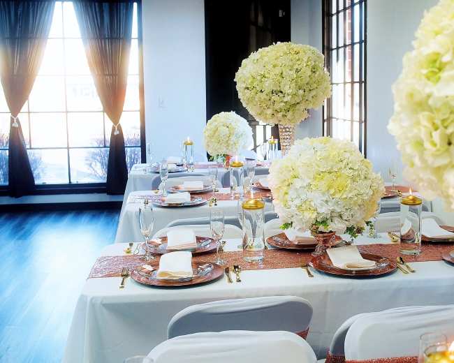 A elegantly set dining area with tables adorned with white floral centerpieces and sparkling table runners, illuminated by natural light through large windows.