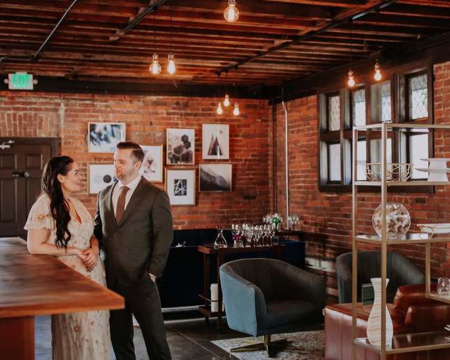 A couple stands together in a stylish venue with exposed brick walls, modern furnishings, and soft overhead lighting.