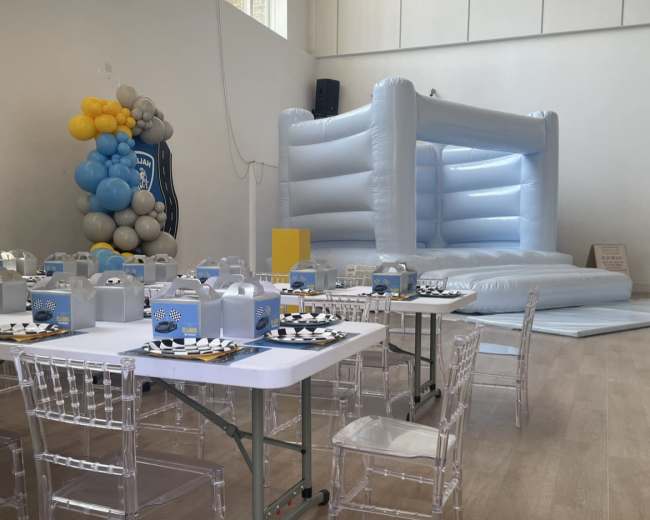 A spacious indoor venue set up for a celebration, featuring a large inflatable bounce house and several tables with clear chairs arranged around them.