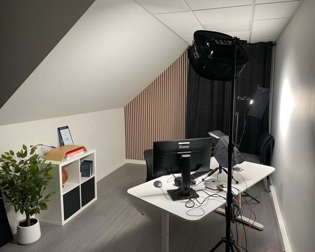 The image shows a modern, minimalistic workspace with a desk, computer setup, and photographic lighting equipment in a well-lit room.