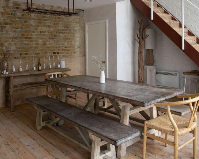 The image shows a rustic dining area featuring a large wooden table with benches, surrounded by a brick wall and a staircase in the background.