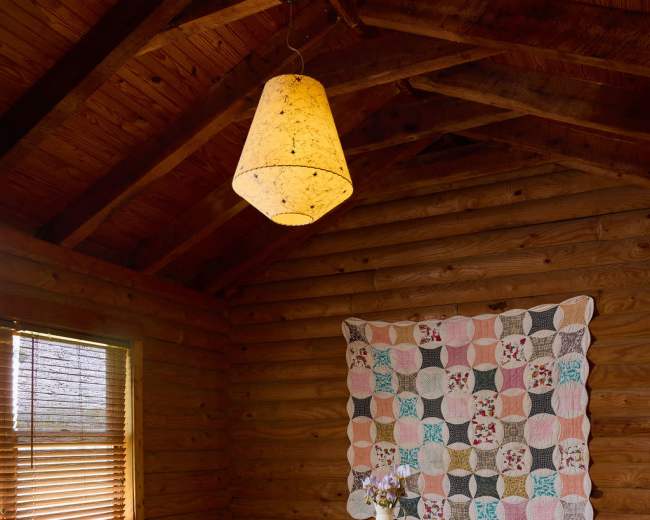 The image shows a rustic interior of a wooden cabin with a table in the center, a quilt hanging on the wall, and a pendant light overhead.