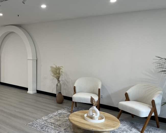 The image shows a modern, minimalist interior space with light-colored walls, a wooden floor, and two chairs arranged around a round table near a decorative archway.