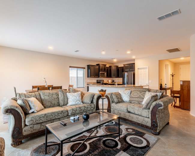 The image shows a spacious living room with two patterned sofas, a coffee table, and a view of an adjacent kitchen area featuring dark cabinets and an open layout.