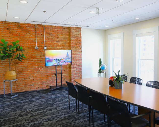 A modern conference room features a long table, black chairs, a brick accent wall, and a television mounted on a stand.