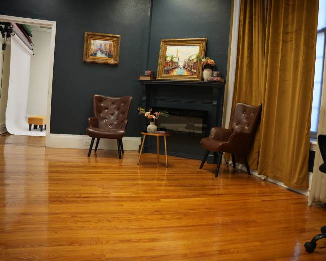 The image shows a room with dark walls, featuring two brown armchairs, a small table, and a decorative fireplace beside a window with yellow curtains.