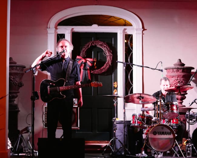 A musician stands with a guitar in front of a microphone while a drummer plays in the background on an outdoor stage.