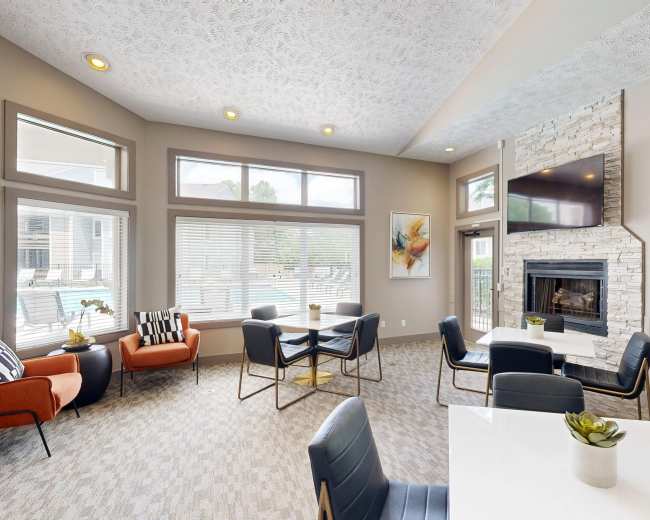 The image shows a modern lounge area with orange and black chairs, a white dining table, a fireplace, and large windows letting in natural light.