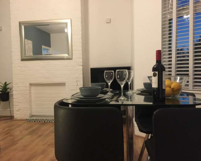 A modern dining area features a round glass table set with two plates, wine glasses, and a bottle of red wine, alongside a bowl of lemons.