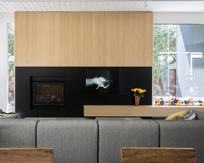 A modern living room features a large gray sofa facing a black wall-mounted television, above a minimalist fireplace, with wooden paneling and a vase of yellow flowers on a light wood console.