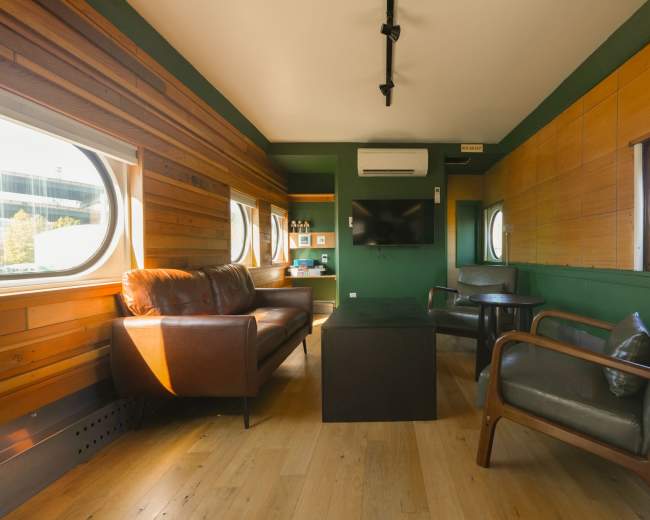 The image shows a modern interior of a room with wooden walls, large round windows, a leather sofa, and a small table in the center.