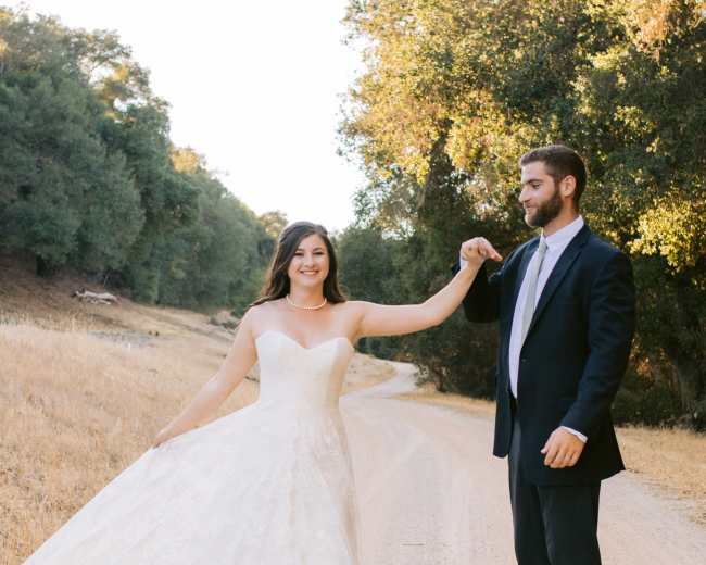 A bride in a white lace gown twirls on a dirt road while holding the hand of a man in a black suit.