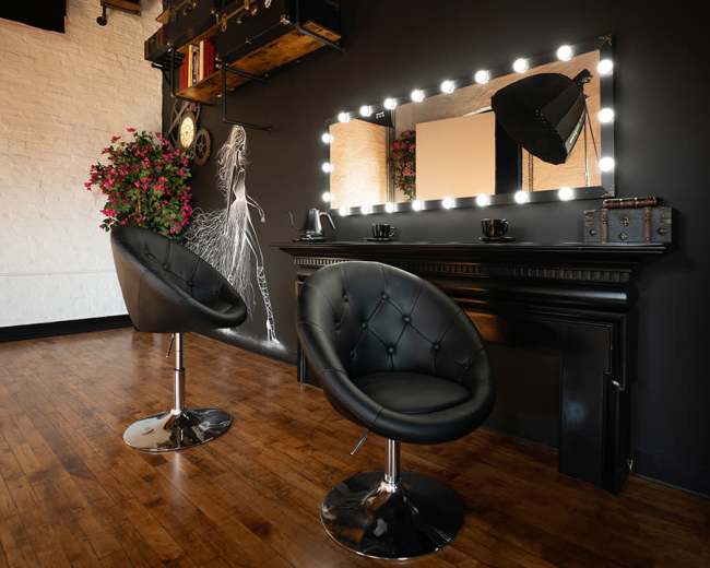 The image shows a minimalistic salon interior with two black, tufted chairs in front of a large lit mirror mounted on a dark wall, accompanied by decorative plants.