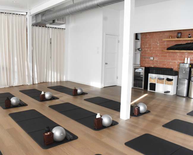 The image shows a minimalist exercise studio with several yoga mats arranged in rows, each featuring a small ball and a bottle on top, along with a kitchenette area in the background.