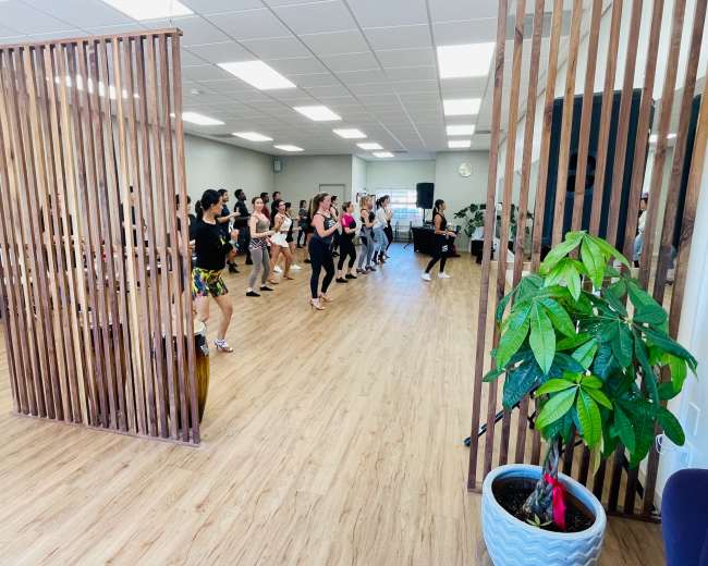 A group of people is participating in a dance class in a spacious studio with wooden flooring and partitioning.