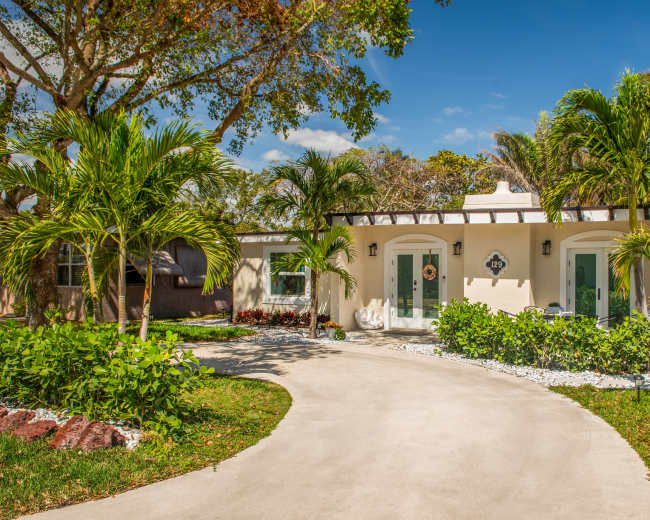 A modern house with a curved driveway is surrounded by lush greenery and palm trees.