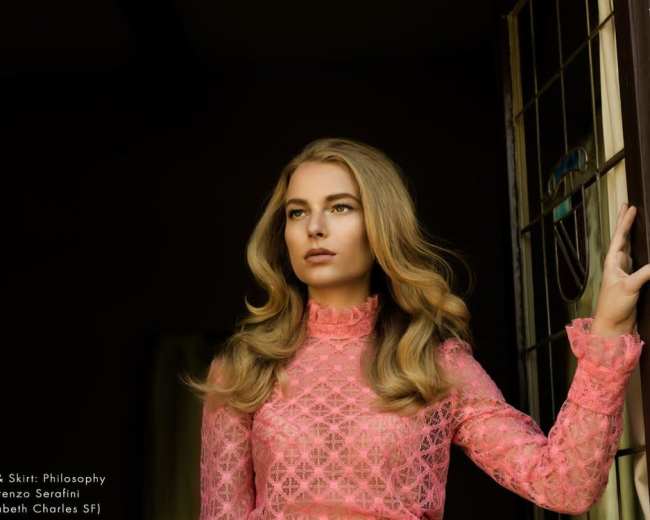 A woman with long hair poses in a pink lace top and skirt near a window.
