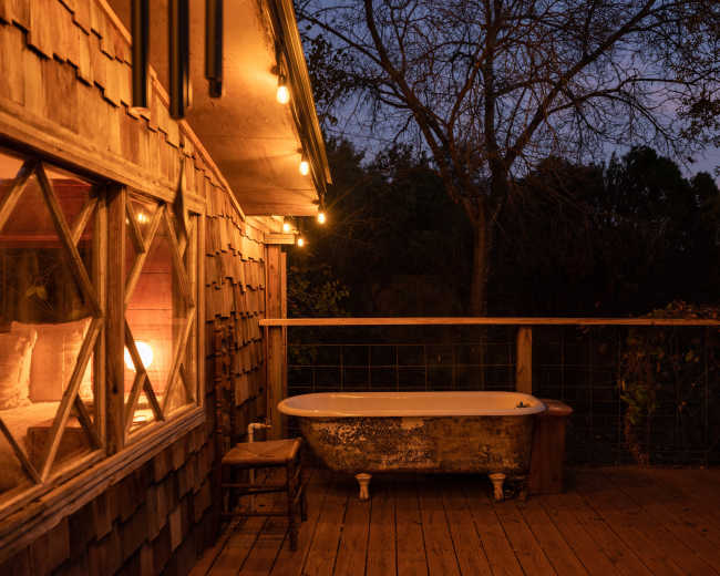 The image shows a vintage clawfoot bathtub on a wooden deck surrounded by trees, with string lights illuminating the scene at dusk.