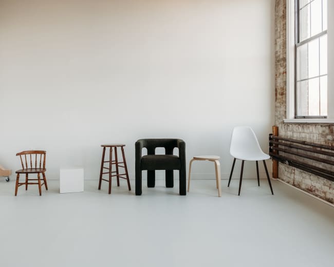 The image shows a row of five different chairs and stools displayed against a plain, light-colored wall in a spacious room.