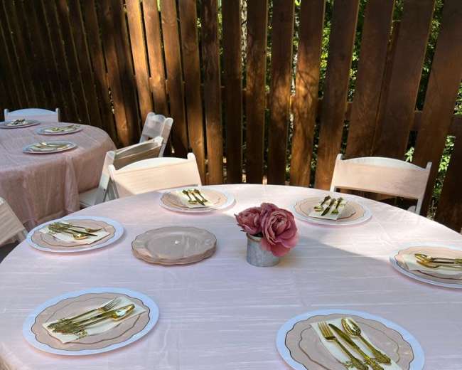 A set of round tables covered with pink tablecloths, each arranged with plates and cutlery, sits against a backdrop of wooden fencing.