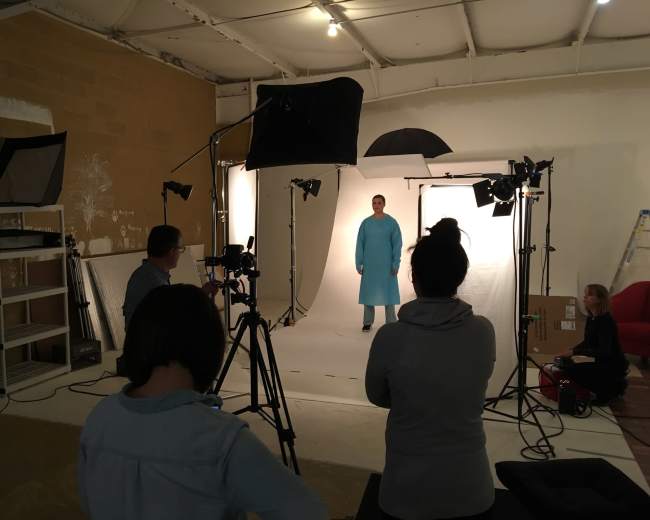 A photoshoot is taking place in a studio, featuring a model in a blue outfit surrounded by multiple cameras and lighting equipment.