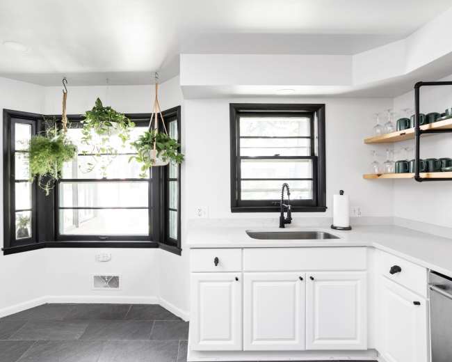 A modern kitchen features a white color scheme with a sink, open shelving, and hanging plants near a large bay window.