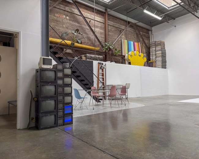 The image shows a spacious industrial-style interior with a staircase, a ladder, a colorful wall hanging, a set of pastel chairs around a table, and a stack of vintage televisions.