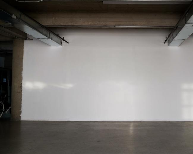 The image shows a large, empty white wall in an industrial-style room with concrete ceilings and floors.