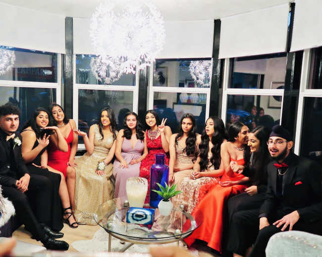 A group of thirteen young adults in formal attire poses together in a decorated living room with large windows and modern lighting.