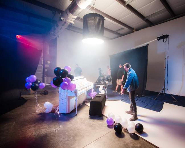 A studio scene features a white piano surrounded by black and purple balloons, with people and equipment in the background.