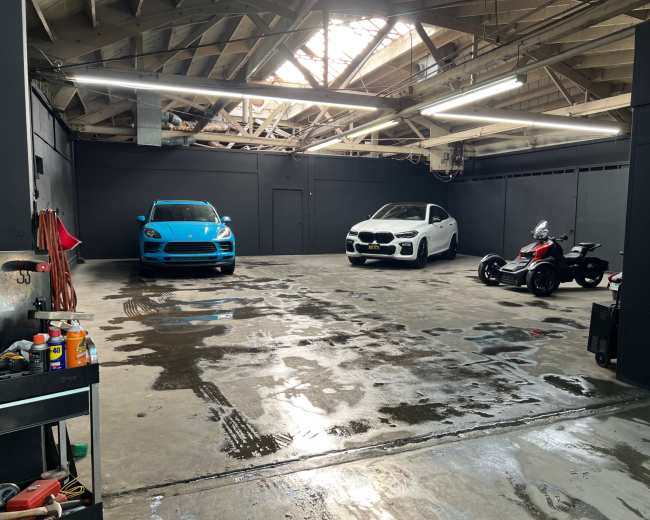 The image shows a spacious garage with a blue car, a white car, and a three-wheeled motorcycle parked on a wet, polished concrete floor under bright overhead lights.