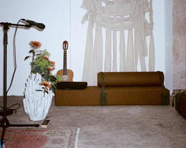 The scene features a minimalist room with a beige fabric wall hanging, a guitar on a stand, a decorative plant, and a candle on the floor.