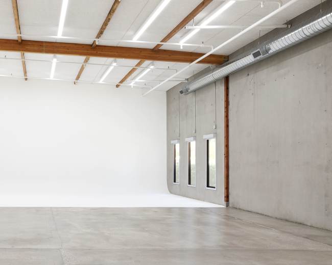 A spacious, minimalist studio with a concrete wall, wooden beams, and bright lighting.