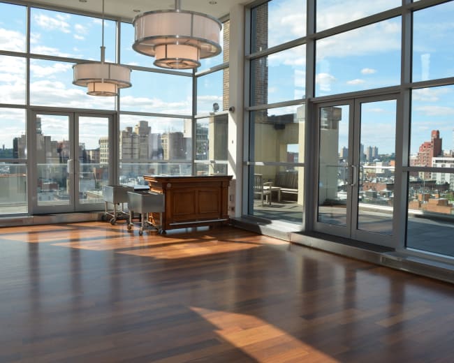 The image shows a spacious, well-lit room with large windows offering a view of a city skyline and wooden flooring.