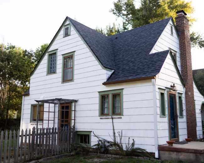 The image shows a two-story, white clapboard house with green trim, a steep roof, and a brick chimney surrounded by trees and a wooden fence.