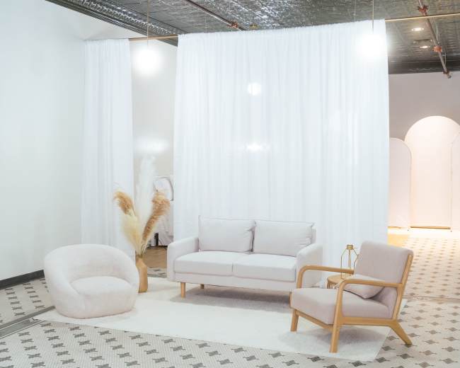 The image features a minimalist interior with a light-colored sofa and armchair, a plush chair, and sheer curtains dividing the space.
