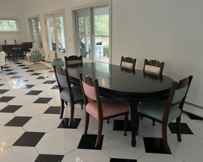 A large black dining table surrounded by eight chairs, some with pink and green upholstery, sits on a black and white checkered tiled floor in a well-lit room with large windows.