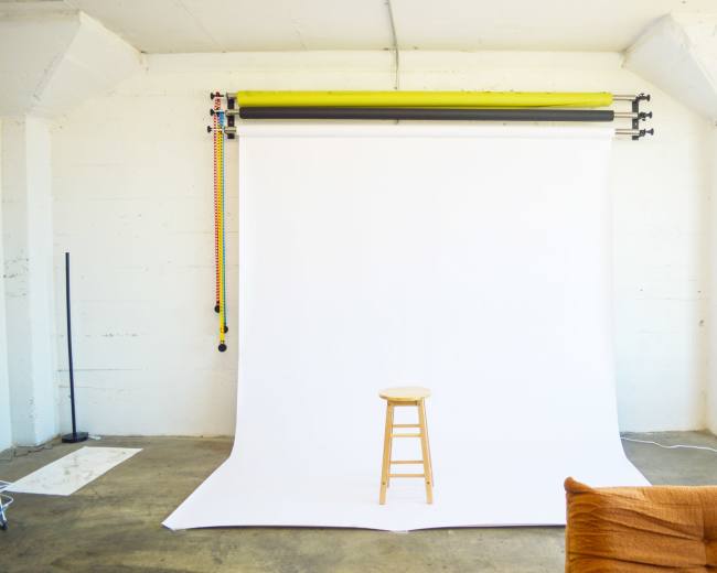 A bare room features a wooden stool in front of a white backdrop supported by a roller system.