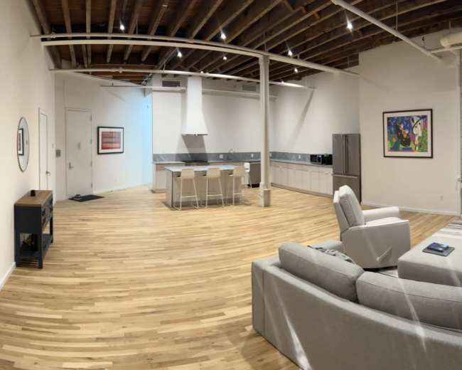 The image shows a spacious, modern interior with a living area featuring a gray sofa, a television, and a kitchen with an island, all surrounded by large windows.