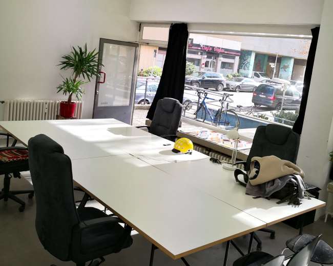 A brightly lit meeting room features a large table surrounded by chairs, with a projector mounted on the ceiling and a window showing street views outside.
