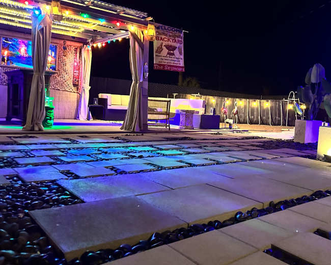 The image shows a brightly lit outdoor backyard area at night, featuring stone pathways, decorative lighting, and lounge furniture.