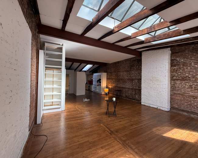 The image shows a spacious, empty loft featuring exposed brick walls, wooden beams, and large skylights that illuminate the wooden floor.