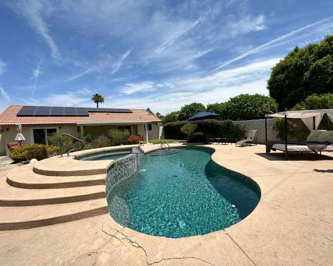 A swimming pool with a spa, surrounded by a patio area with lounge chairs and an umbrella, and a house with solar panels in the background.